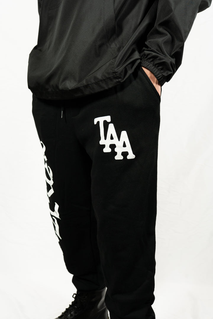 A person wearing black The Amity Affliction fleece sweatpants with "TAA" in large white letters on the left thigh and a black jacket stands against a white background, showing only the lower half of their body.