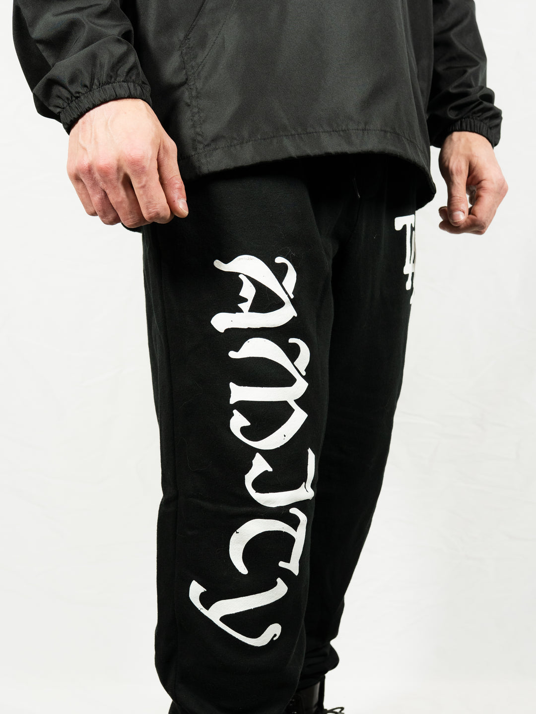 A person wearing black midweight fleece sweatpants from The Amity Affliction with ANXIETY in large white gothic letters down the left leg, paired with a black jacket, standing against a plain white background.