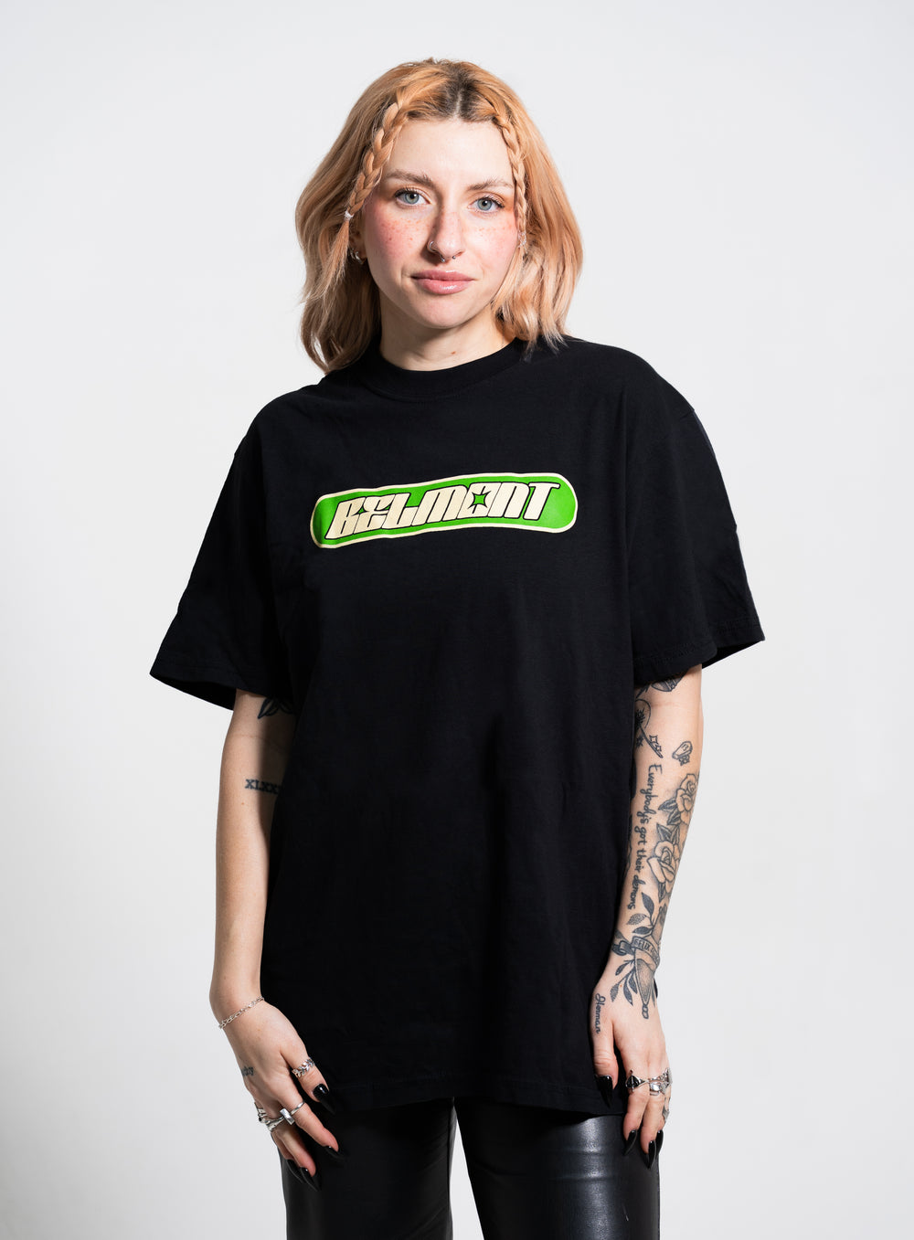 A person with light brown, wavy hair and tattoos on both arms wears black pants and a black Belmont t-shirt with a green and white logo, standing against a plain light background.