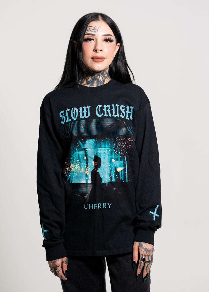 A woman with long black hair, face and neck tattoos, and black clothes stands against a plain background. She wears a Slow Crush Cherry t-shirt with blue graphics on Gildan apparel, holding her hands in front of her.