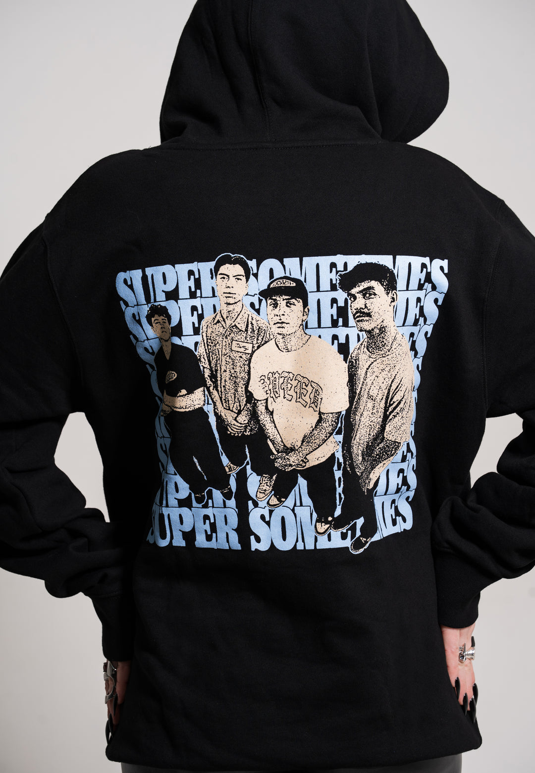 A person wears a black Super Sometimes hoodie featuring four illustrated young men on the back, with "SUPER SOMETIMES" repeated in blue and white behind them. Their hands are visible at their sides.