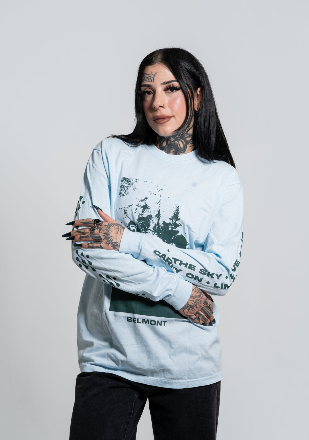 A woman with long dark hair, face and neck tattoos, and black nail polish stands with arms crossed, wearing a light blue Belmont graphic long-sleeve shirt and black pants against a plain light background.