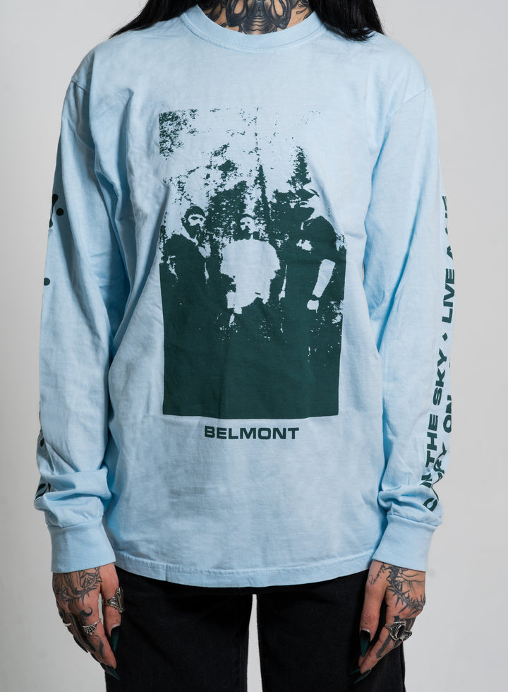 A tattooed person wears a Belmont light blue long-sleeve shirt with a dark band graphic and BELMONT printed on the front, plus text along the sleeves.