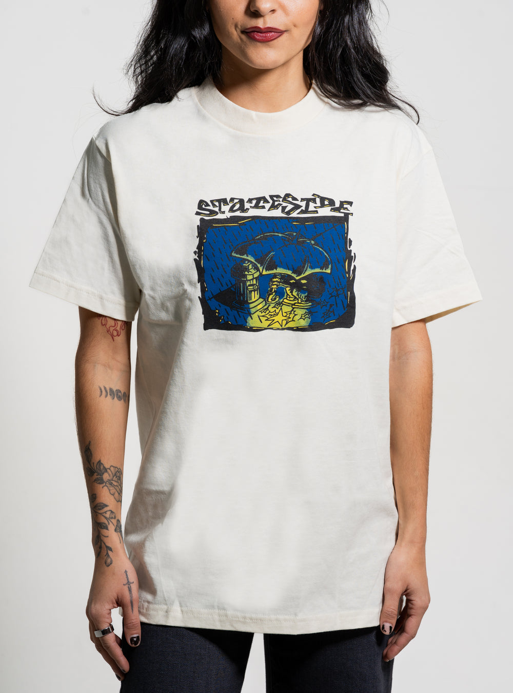 A woman with long dark hair and visible arm tattoos stands against a plain background, wearing a white Stateside T-shirt featuring a colorful rectangular graphic and the word "Statefire" on the chest.