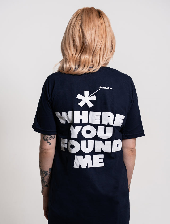 A person with shoulder-length blonde hair stands facing away, wearing a Stateside black T-shirt with “* WHERE YOU FOUND ME” in bold white letters, and has visible tattoos on their arms.