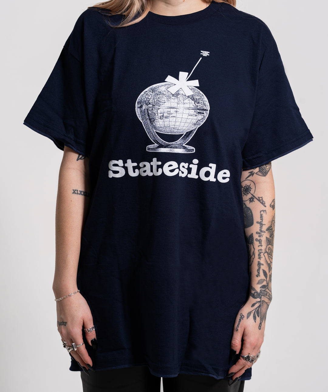 A person with tattooed arms wears a loose dark blue Stateside T-shirt featuring a globe graphic with a large asterisk and "Stateside" printed below in bold white letters.