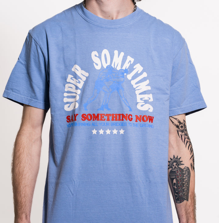 A person wears a light blue Super Sometimes Comfort Colors t-shirt with white and red "SAY SOMETHING NOW" text, subtle graphic, and has a large sun and moon tattoo on their right arm.