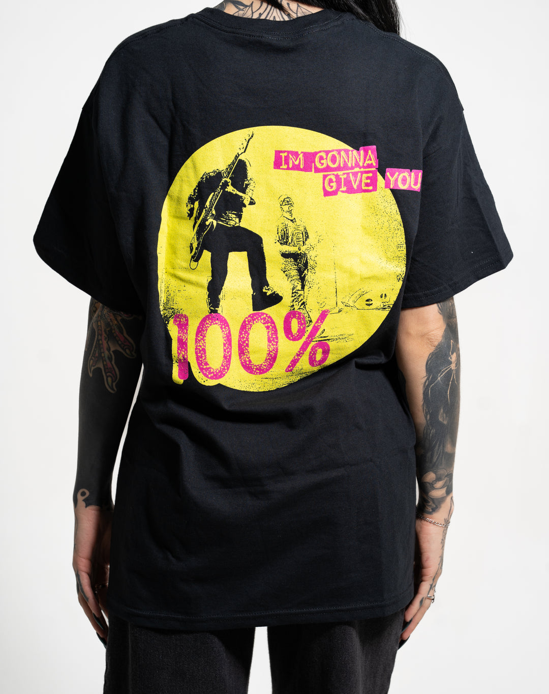 A person with tattoos wears a black New Found Glory tee featuring a yellow circle with two figures, pink text "IM GONNA GIVE YOU" on the front, and bold pink-yellow "100%" on the back.