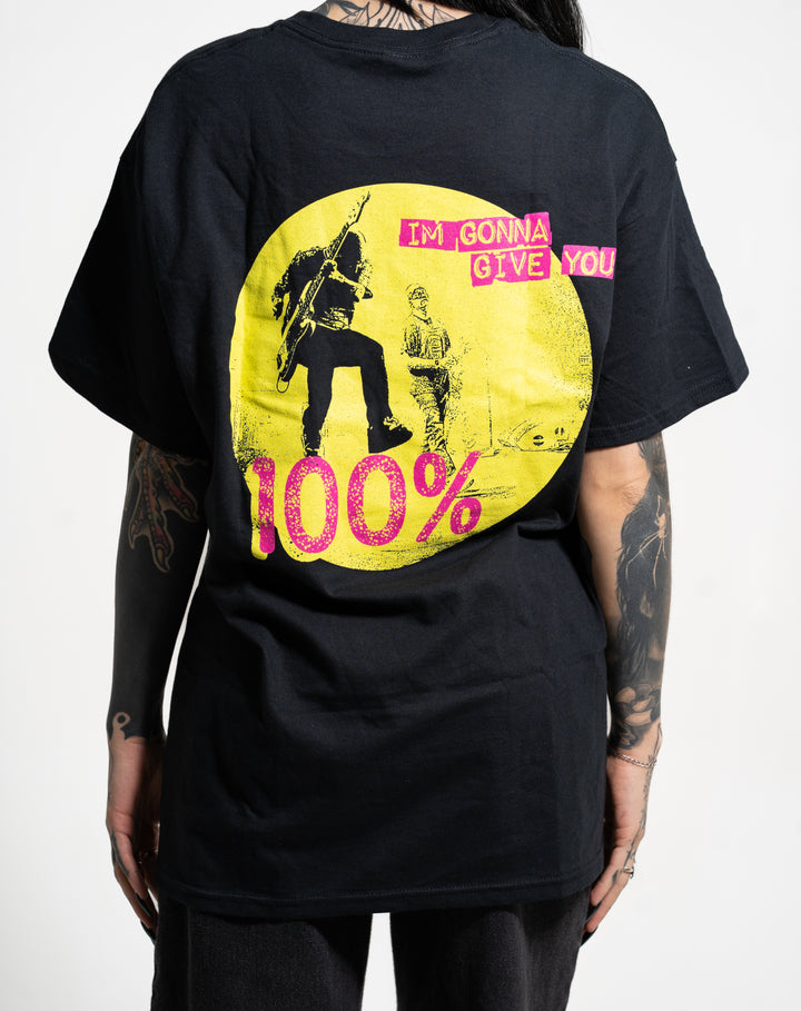 A person with tattoos wears a black New Found Glory tee featuring a yellow circle with two figures, pink text "IM GONNA GIVE YOU" on the front, and bold pink-yellow "100%" on the back.