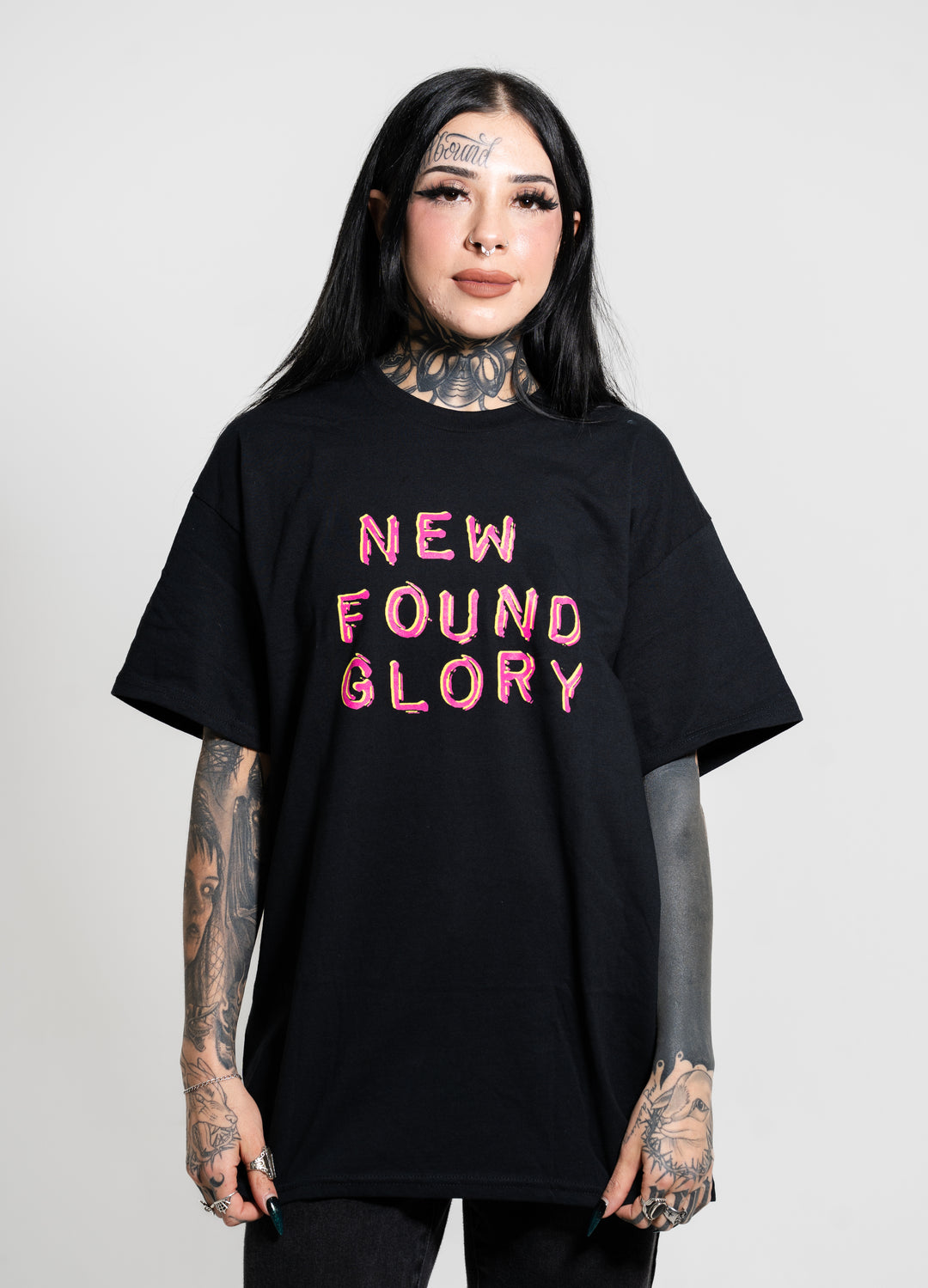 A person with long dark hair and tattoos stands against a plain background, wearing a black New Found Glory t-shirt with the band name in pink letters on the front.