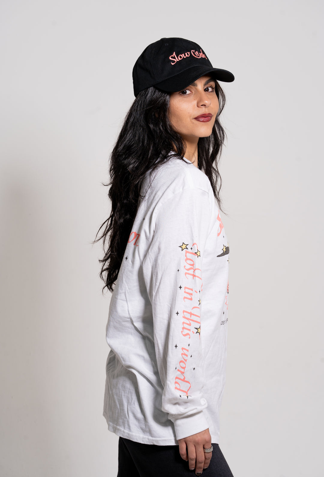 A woman with long dark hair models Slow Crush apparel: a black dad hat, a white long-sleeve shirt with sleeve graphics, and black pants. She stands sideways, facing the camera against a plain white background.