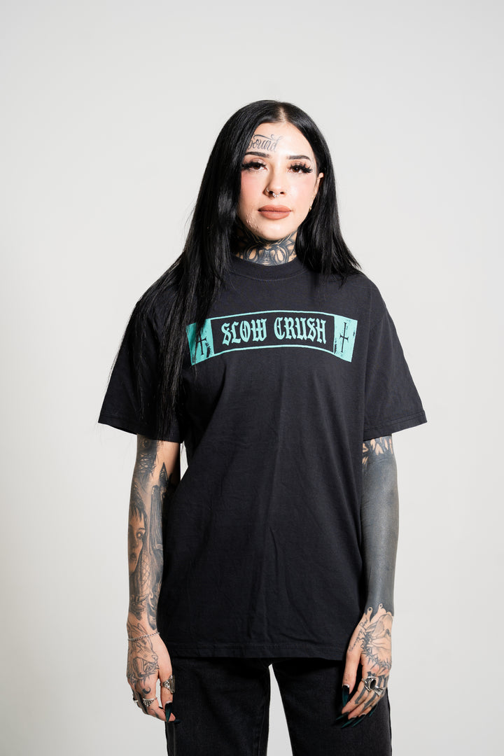 A person with long black hair and visible tattoos stands facing forward against a white background, wearing a black Slow Crush t-shirt with teal "SLOW CRUSH" lettering and black pants.