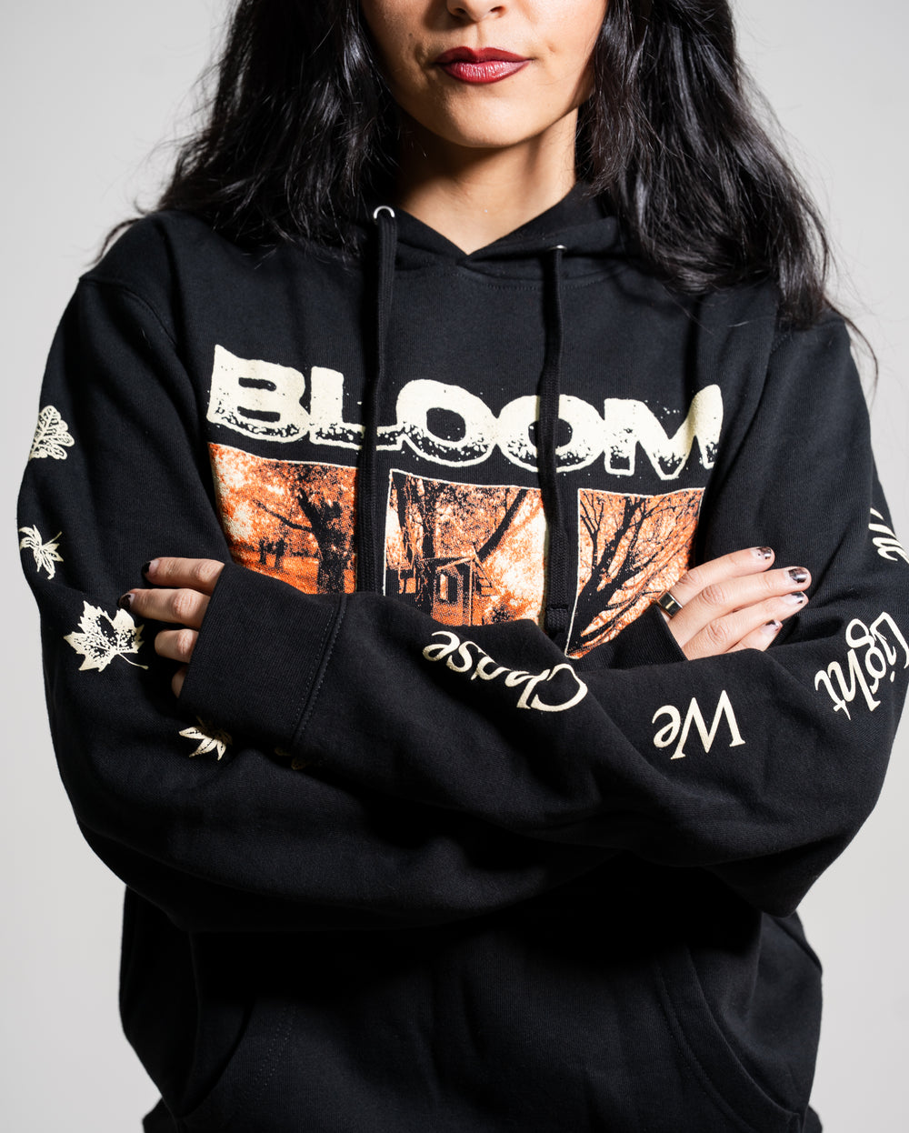 A woman with long dark hair and red lipstick wears a black Bloom hoodie featuring “BLOOM” and an autumn scene on the front. Her arms are crossed, showing decorative words and leaves on the sleeves.
