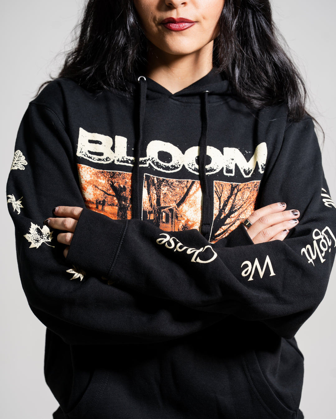 A woman with long dark hair and red lipstick wears a black Bloom hoodie featuring “BLOOM” and an autumn scene on the front. Her arms are crossed, showing decorative words and leaves on the sleeves.