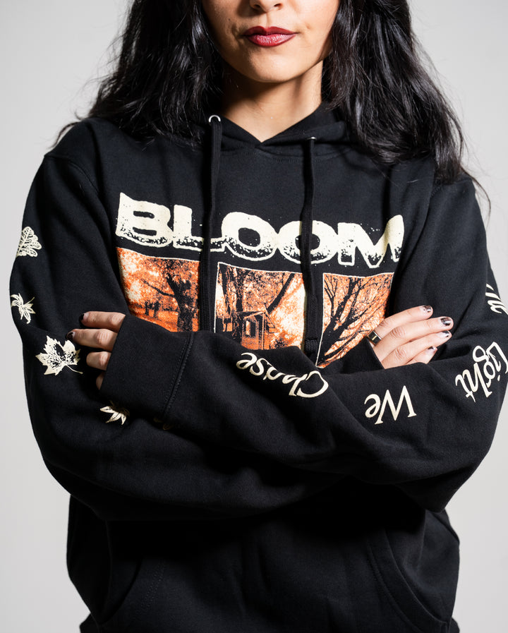 A woman with long dark hair and red lipstick wears a black Bloom hoodie featuring “BLOOM” and an autumn scene on the front. Her arms are crossed, showing decorative words and leaves on the sleeves.
