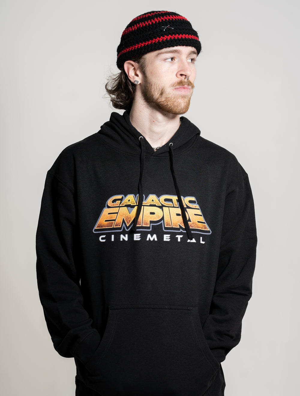 A man with light skin, a beard, and long hair wears a black Galactic Empire hoodie with "Cinemetal" text and a black and red knit hat. He looks to the side against a plain, light background.