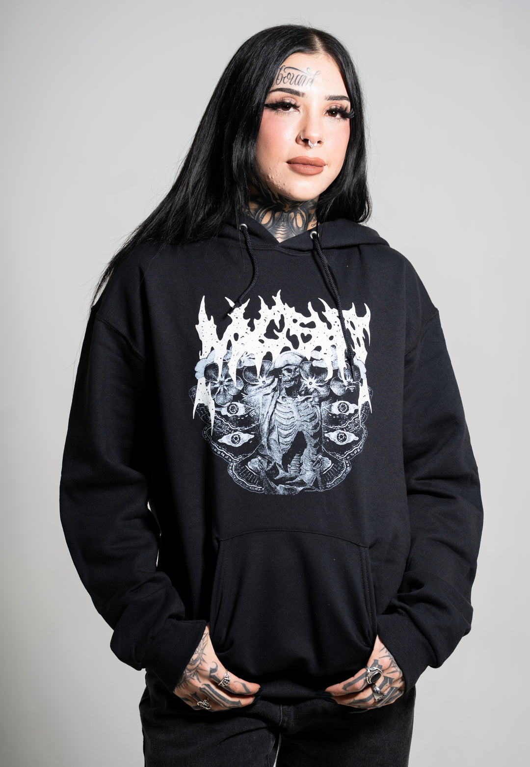 A person with long black hair, tattoos, and bold makeup wears a Mugshot black hoodie featuring a white, intricate gothic graphic. They stand against a plain light gray background, hands in pockets and smiling.