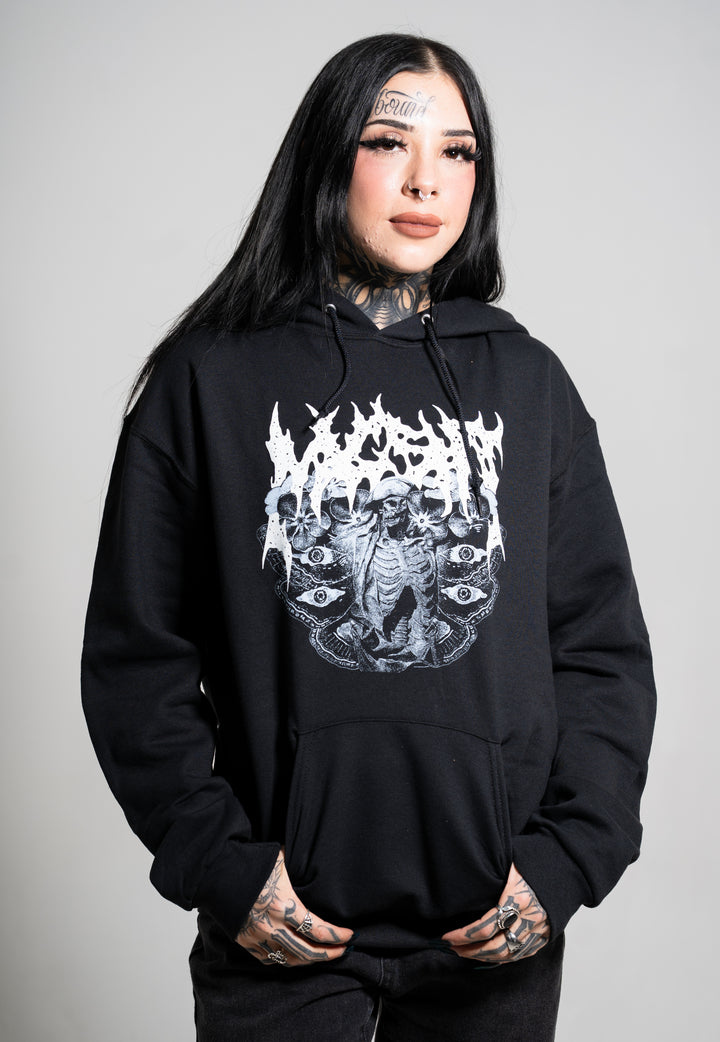 A person with long black hair, tattoos, and bold makeup wears a Mugshot black hoodie featuring a white, intricate gothic graphic. They stand against a plain light gray background, hands in pockets and smiling.