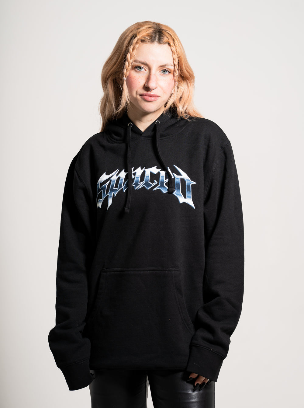 A young woman with light reddish-blonde braided hair wears a black Spaced hoodie featuring bold, stylized graphic text on the front. She stands against a plain light background, looking at the camera with a slight smile.