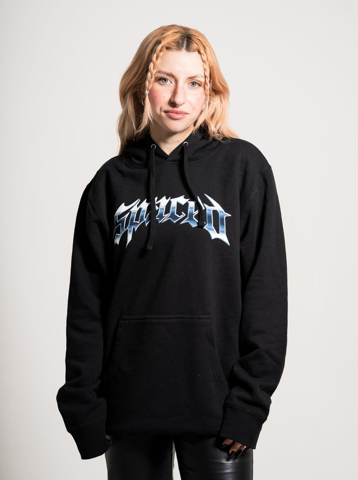 A young woman with light reddish-blonde braided hair wears a black Spaced hoodie featuring bold, stylized graphic text on the front. She stands against a plain light background, looking at the camera with a slight smile.