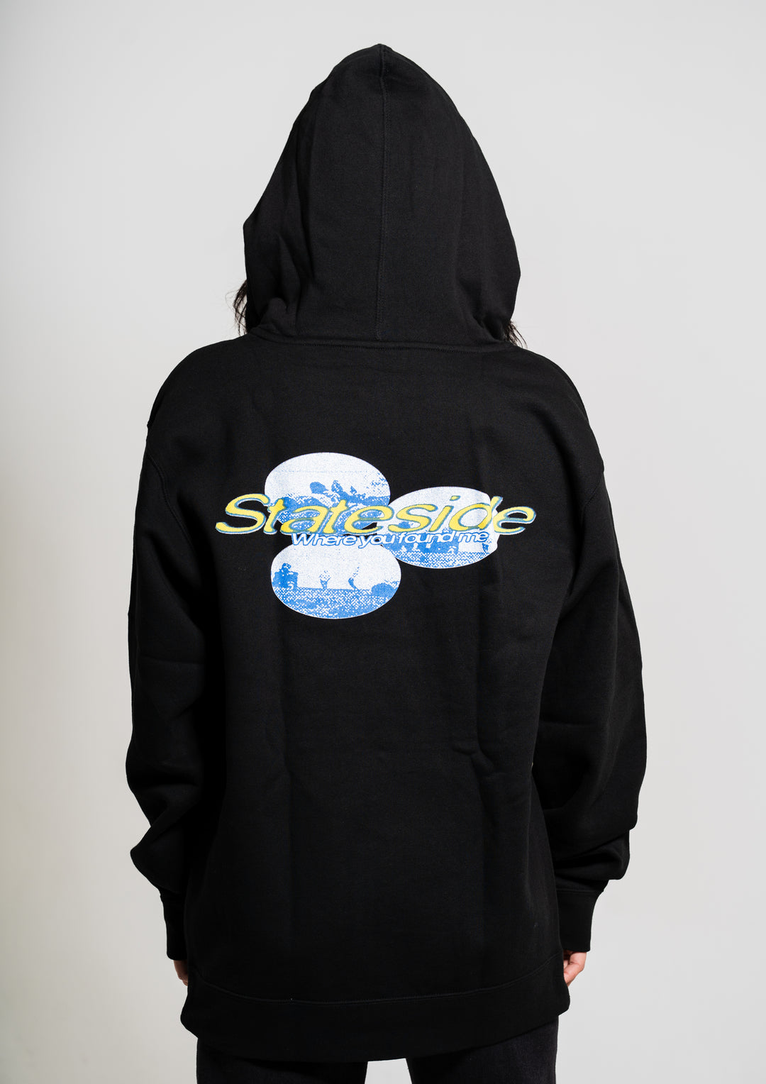 Someone wearing a black Stateside hoodie with the hood up. The back features two blue and white circles, "Stateside" in yellow, and "Neighborhood" in blue underneath.