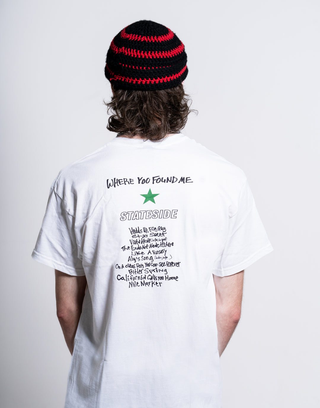 A person with long hair, seen from behind against a white background, wears a black and red knitted beanie and a Stateside white T-shirt featuring text and a green star on the back.