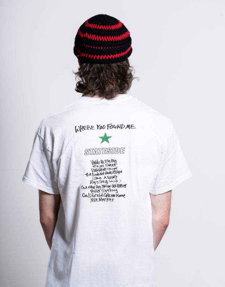A person with long hair, seen from behind against a white background, wears a black and red knitted beanie and a Stateside white T-shirt featuring text and a green star on the back.