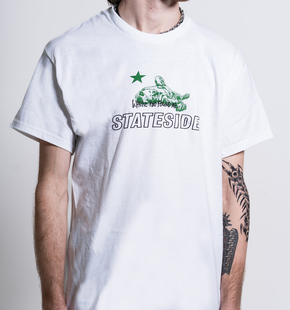 A person with a tattooed left arm stands against a plain background wearing the Stateside white T-shirt featuring a green star, turtle graphic, and the text “WHERE YOU FOUND ME STATESIDE.”.