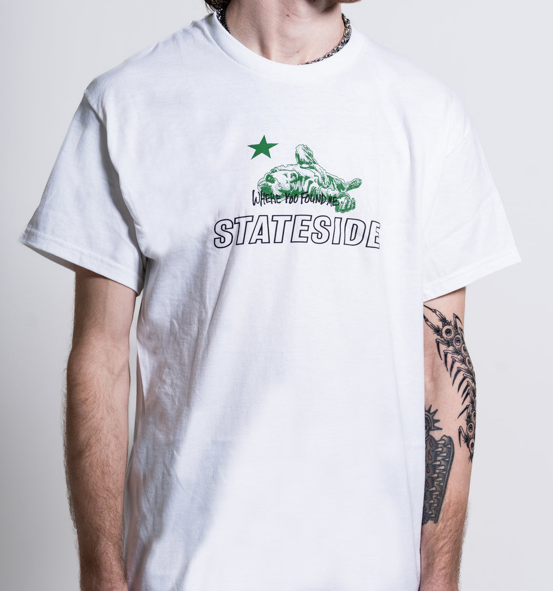 A person with a tattooed left arm stands against a plain background wearing the Stateside white T-shirt featuring a green star, turtle graphic, and the text “WHERE YOU FOUND ME STATESIDE.”.