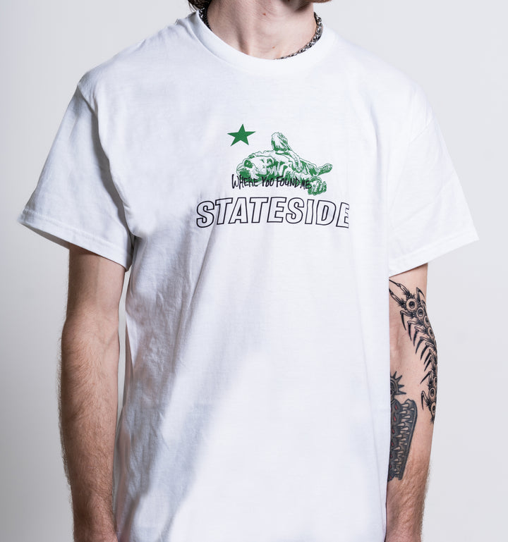 A person with a tattooed left arm stands against a plain background wearing the Stateside white T-shirt featuring a green star, turtle graphic, and the text “WHERE YOU FOUND ME STATESIDE.”.