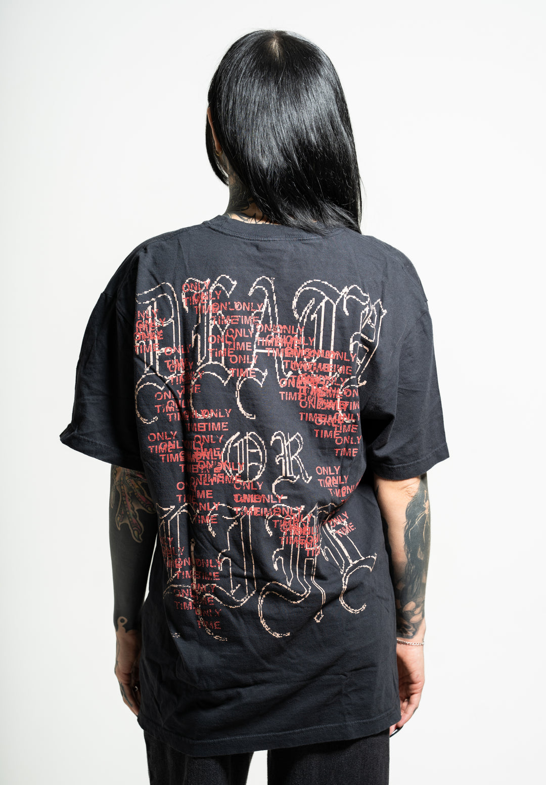 A person with long dark hair faces away, wearing a black Soulkeeper t-shirt with bold Gothic white lettering and repeated red "ONLY TIME WILL TELL" text across the back; tattoos adorn both arms.