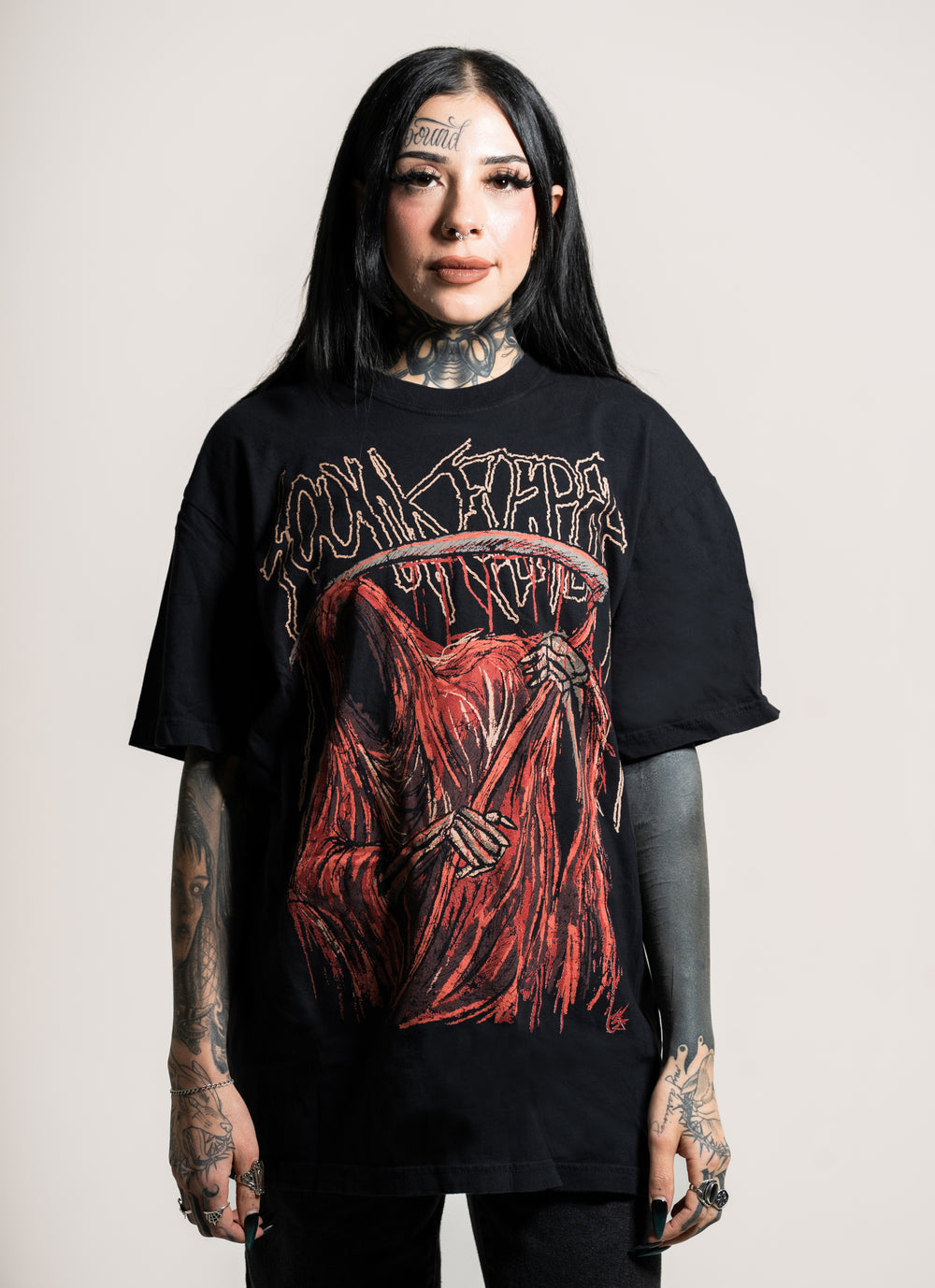 A woman with long black hair and tattoos faces the camera wearing a black Soulkeeper Comfort Colors t-shirt with a red grim reaper graphic and dark pants, standing against a plain light background.