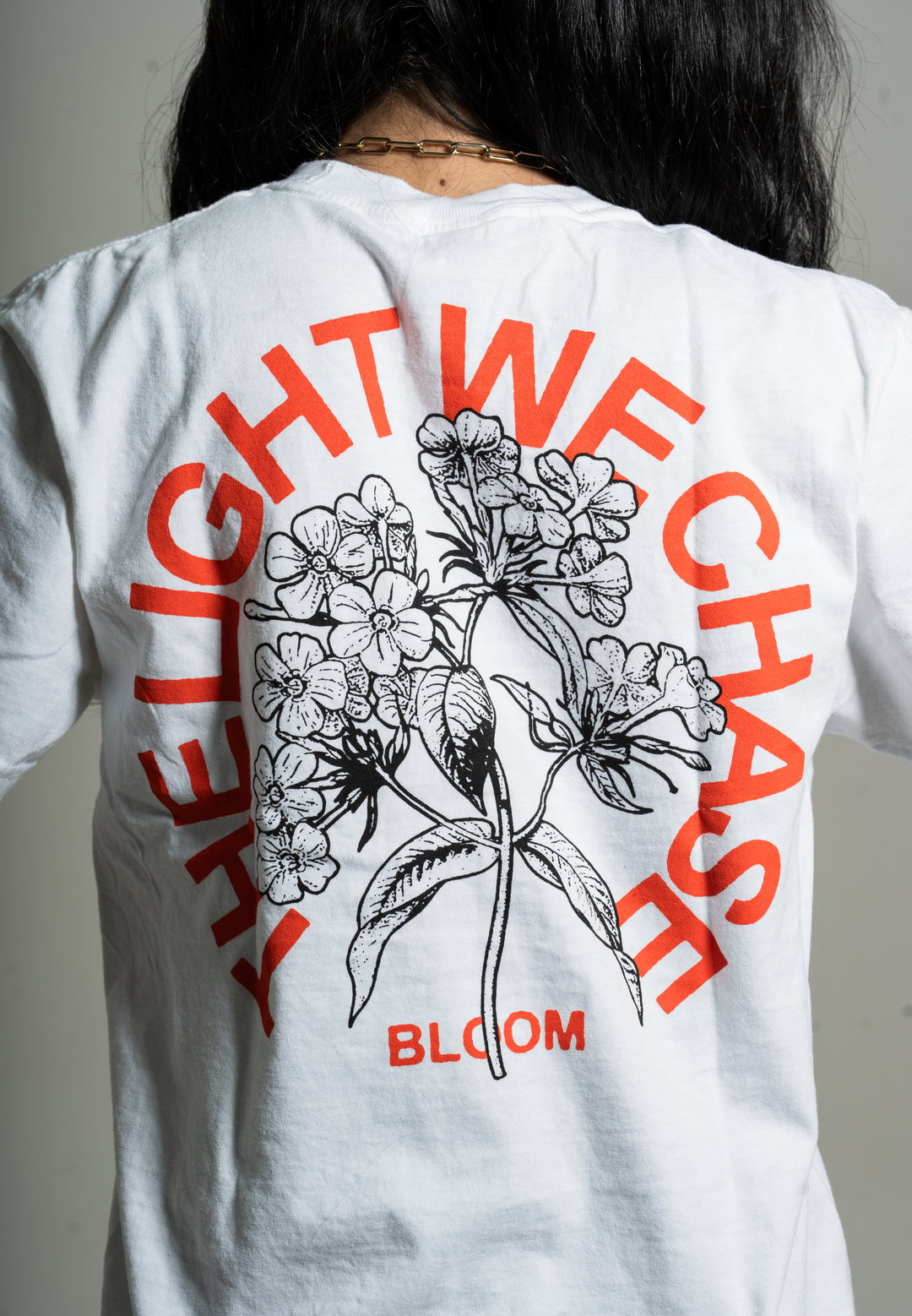 Someone wears a white Bloom shirt featuring a large black floral illustration on the back. Red text arcs around the flowers, saying “THE LIGHT WE CHASE,” with “BLOOM” printed below in red letters.
