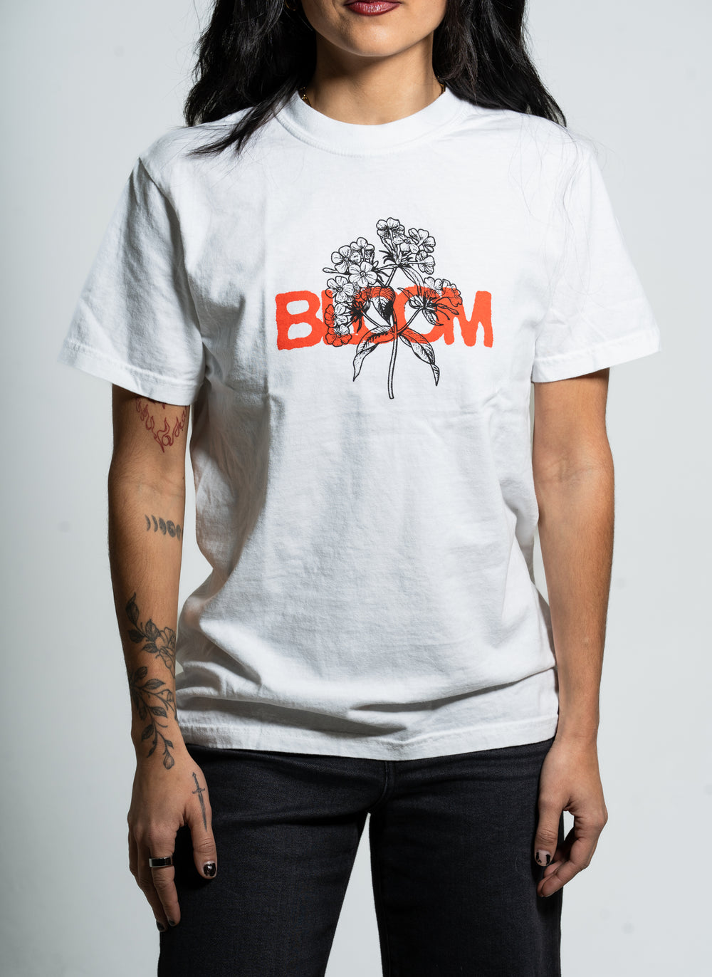 A person with tattooed arms wears a Bloom brand white t-shirt featuring a black floral line drawing and the word "BLOOM" in bold orange letters across the chest, standing against a plain background.
