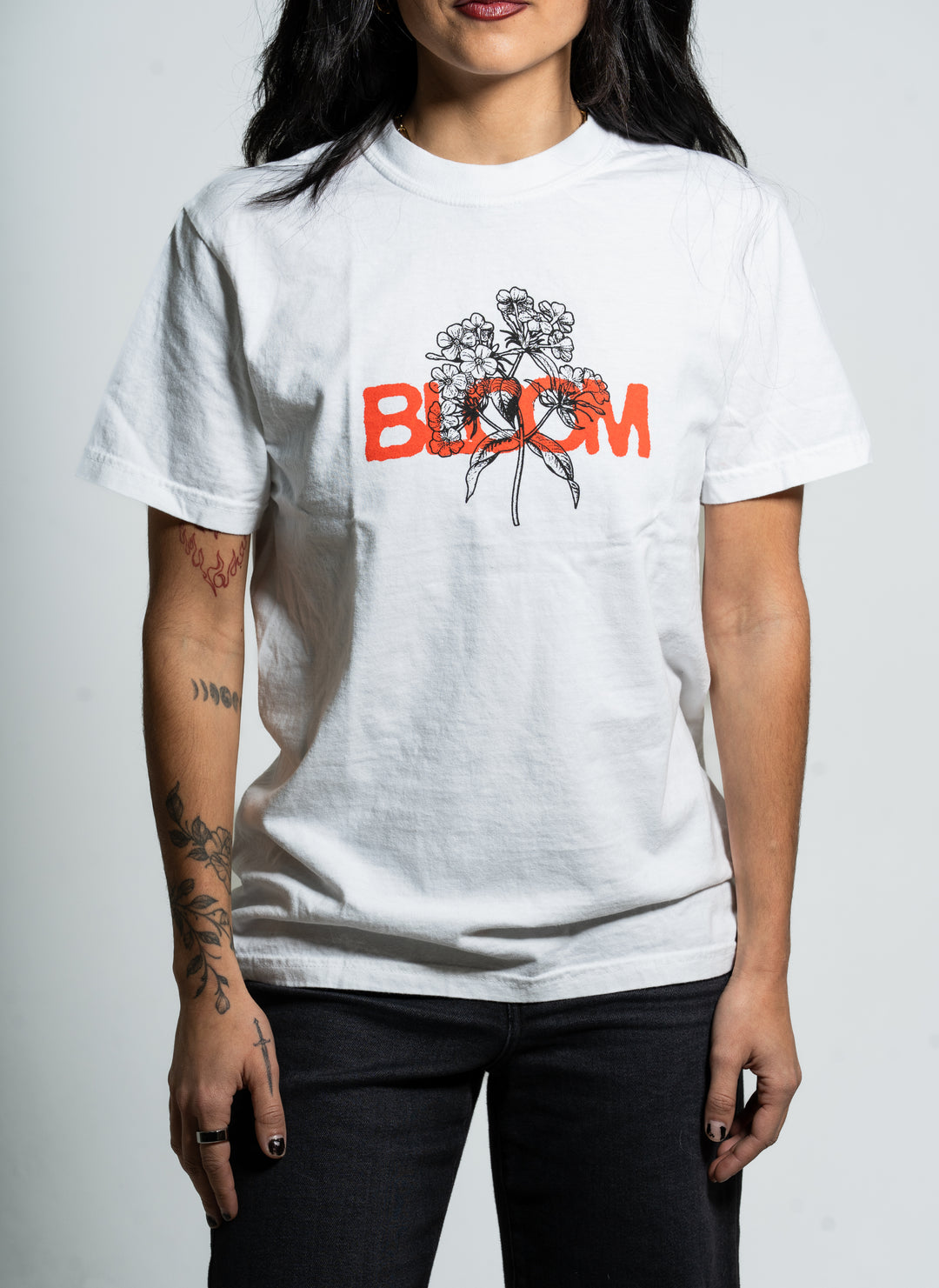 A person with tattooed arms wears a Bloom brand white t-shirt featuring a black floral line drawing and the word "BLOOM" in bold orange letters across the chest, standing against a plain background.
