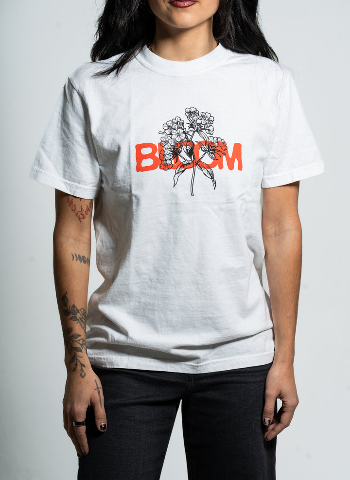 A person with tattooed arms wears a Bloom brand white t-shirt featuring a black floral line drawing and the word "BLOOM" in bold orange letters across the chest, standing against a plain background.