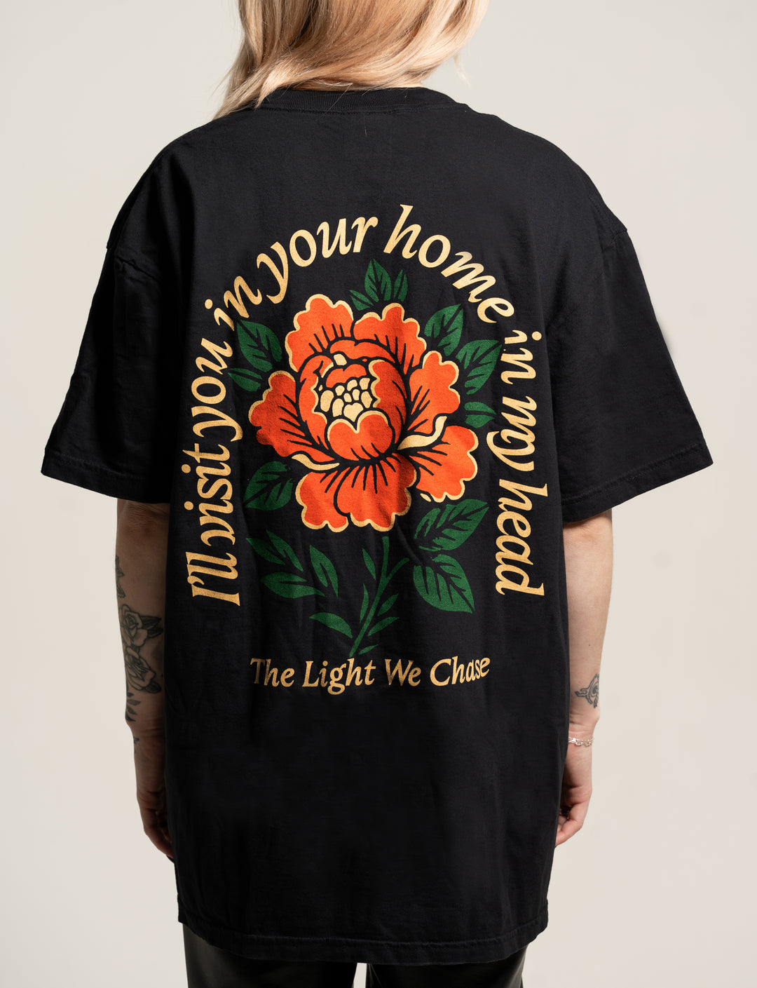A person wears a black Bloom t-shirt featuring a large orange flower with green leaves graphic on the back, surrounded by the text: "I'll visit you in your home, in my head. The Light We Chase.