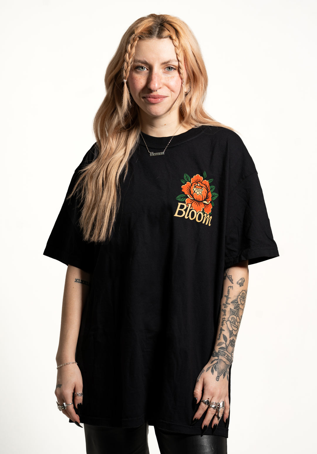 A woman with long, wavy blonde hair and tattooed arms stands against a white background, wearing a loose black Bloom T-shirt featuring an orange flower and the word "Bloom." She smiles slightly.