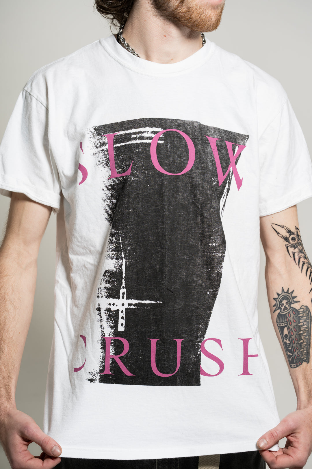 A person wears a white Slow Crush Comfort Colors t-shirt with a bold black abstract graphic and "SLOW CRUSH" in pink. Their tattooed arm, partial face, and a chain necklace are also visible.