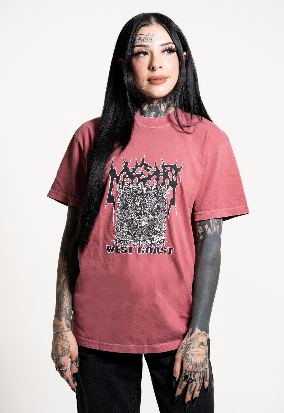 A tattooed woman with long black hair wears a maroon Mugshot graphic T-shirt reading "WEST COAST" in bold, jagged print. She stands against a plain white background, gazing off to the side as if absorbed in thought.