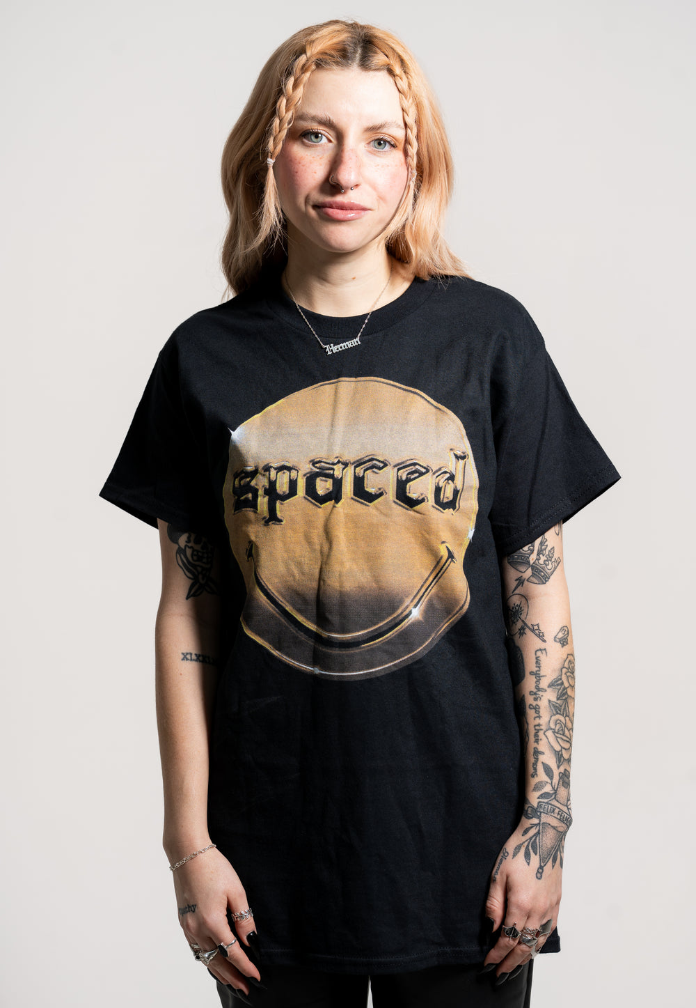 A person with light brown hair and arm tattoos faces the camera, wearing a black Spaced t-shirt featuring the brand name in bold letters within a circular design. The background is plain and light-colored.
