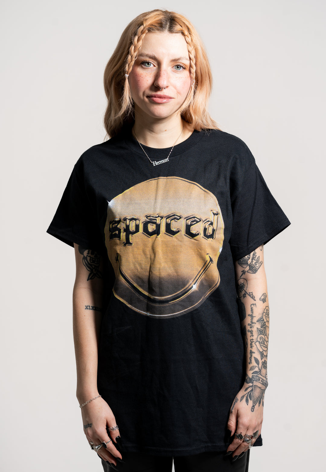 A person with light brown hair and arm tattoos faces the camera, wearing a black Spaced t-shirt featuring the brand name in bold letters within a circular design. The background is plain and light-colored.