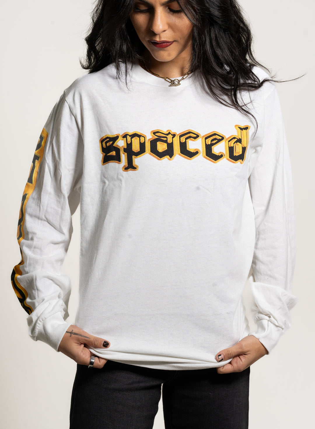 A person with long dark hair wears a white Spaced long-sleeve shirt with bold yellow and black logo on the front and printed sleeve designs, looking down while holding the shirt hem.