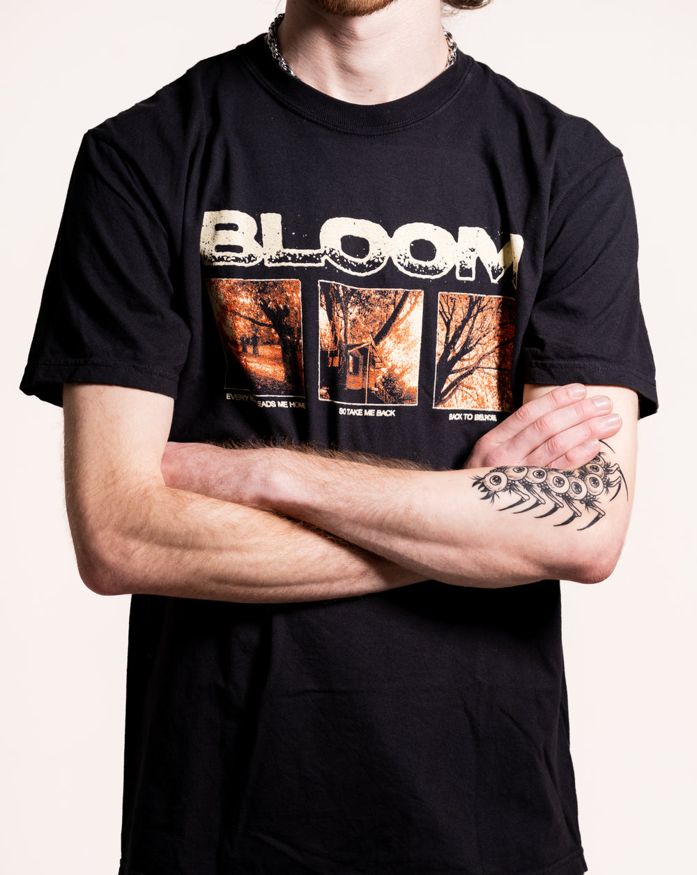 A person with crossed arms wears a black Bloom T-shirt with "BLOOM" above three autumn images, a chain necklace, and a black fishbone tattoo on their left forearm. The background is plain white.