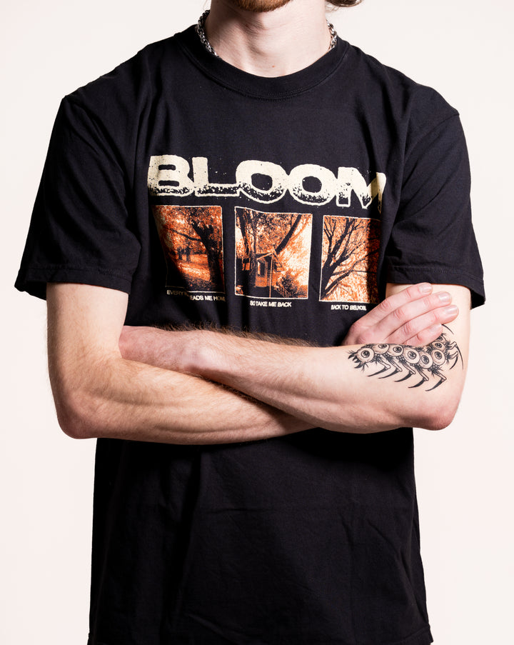 A person with crossed arms wears a black Bloom T-shirt with "BLOOM" above three autumn images, a chain necklace, and a black fishbone tattoo on their left forearm. The background is plain white.