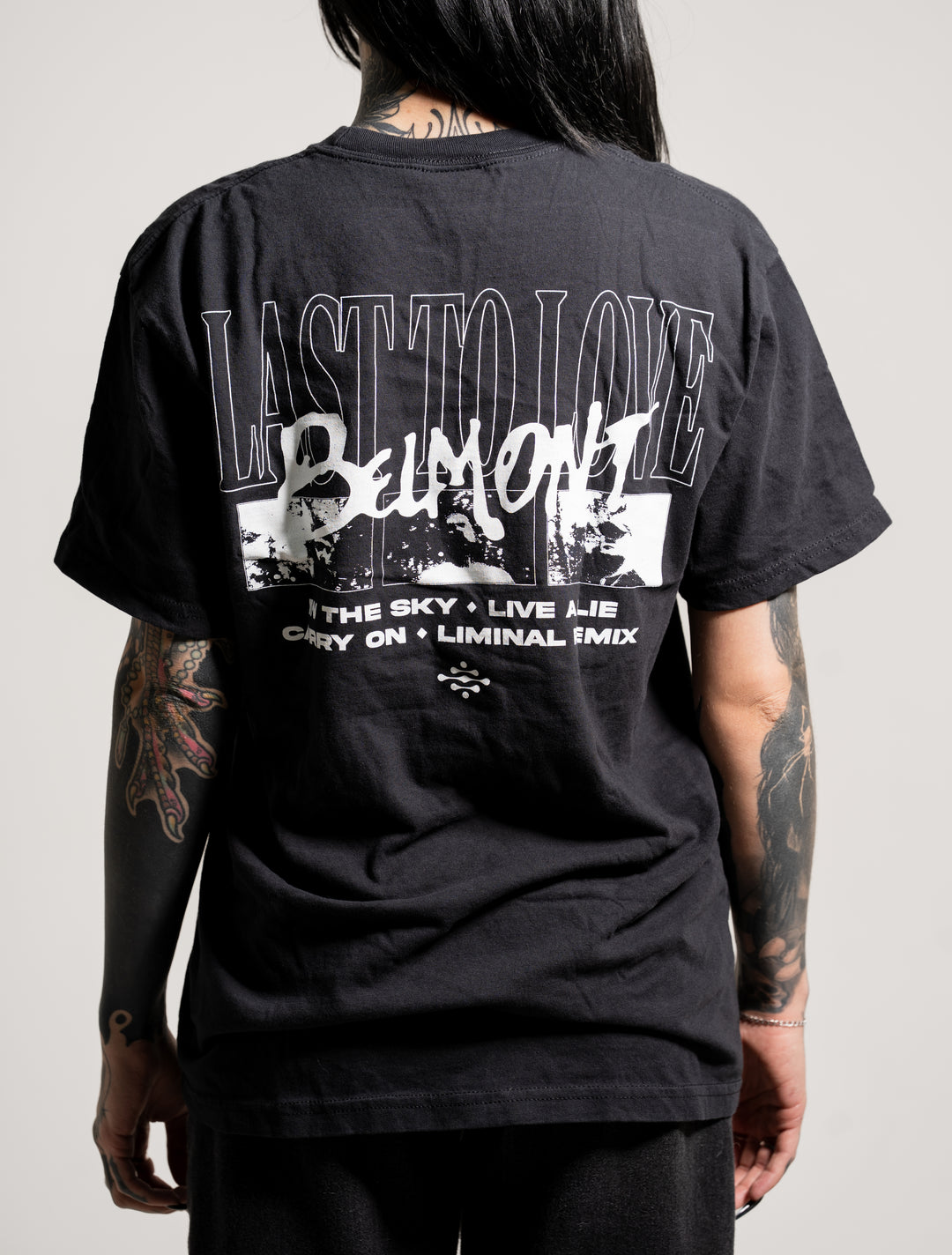 A person with tattooed arms faces away, wearing a black Belmont T-shirt featuring white graphics and the text: LAST TO LOVE BELMONT THE SKY • LIVE A LIE CRY ON • LIMINAL FMIX. The background is plain and neutral.