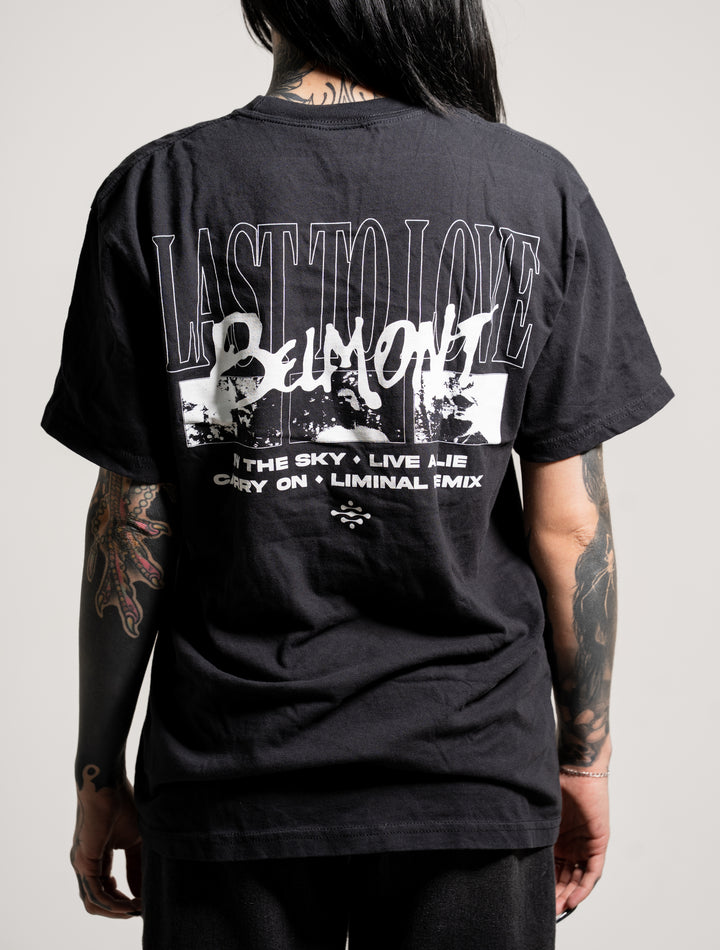 A person with tattooed arms faces away, wearing a black Belmont T-shirt featuring white graphics and the text: LAST TO LOVE BELMONT THE SKY • LIVE A LIE CRY ON • LIMINAL FMIX. The background is plain and neutral.