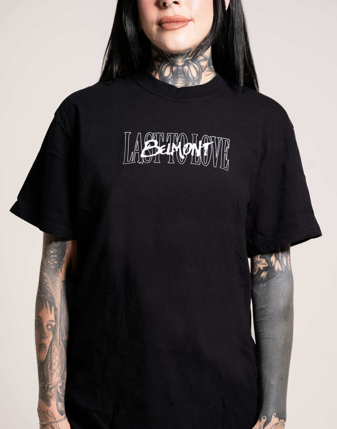 A person with long dark hair and tattoos on their neck and arms wears a black Belmont T-shirt that reads LAST TO LOVE, with SORROW scribbled over TO in white text.