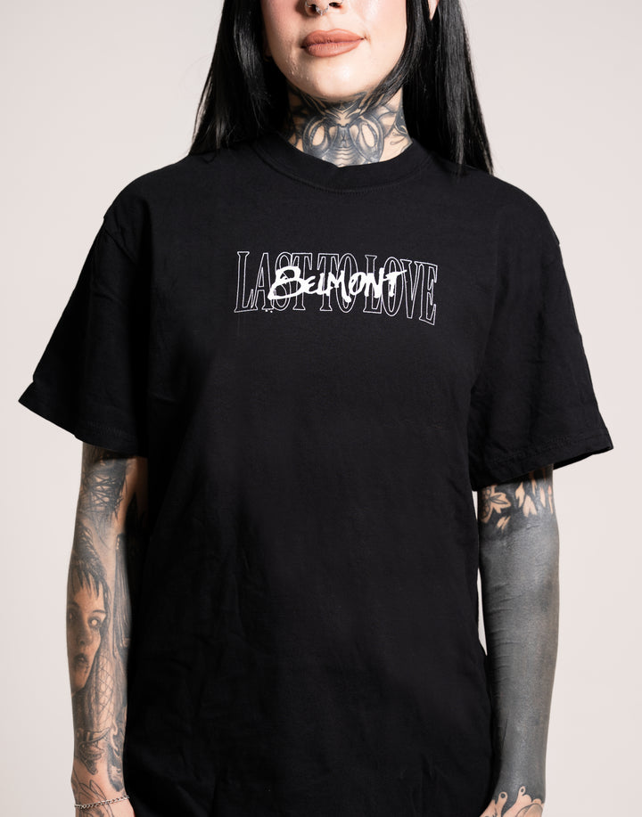 A person with long dark hair and tattoos on their neck and arms wears a black Belmont T-shirt that reads LAST TO LOVE, with SORROW scribbled over TO in white text.