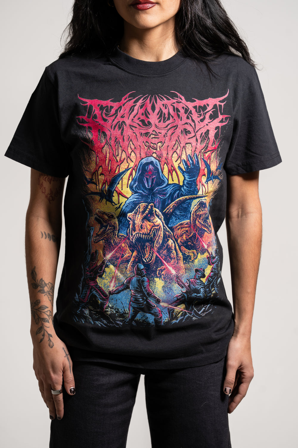 A person with long dark hair and tattooed arms wears a Galactic Empire black T-shirt with colorful artwork of a hooded figure and three fierce dinosaurs in a dynamic, fiery scene. The shirt displays a stylized band logo.